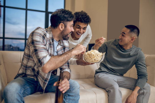 Cheerful guy with a bowl of popcorn in the hand leaning towards the guests seated on the sofa - Photo, image