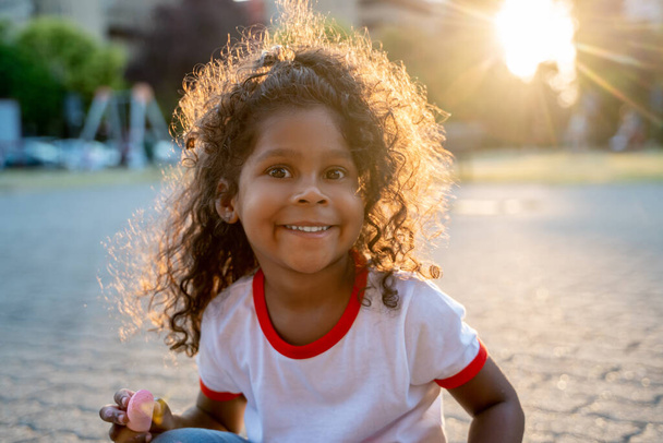 Criança feminina alegre com um pedaço de giz colorido na mão sentada no pavimento olhando para frente - Foto, Imagem