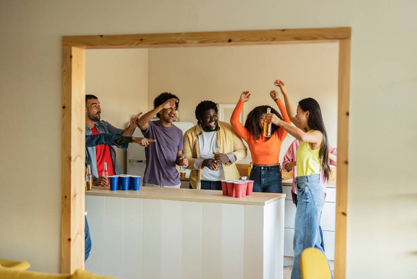 Group of multiracial friends with raised arms gathered in a kitchen while serving alcoholic beers to celebrate a party. - Photo, Image