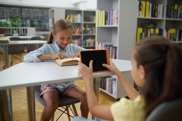 Modern school. Girls spending time in the library, one of them using a smartphone - Foto, Bild