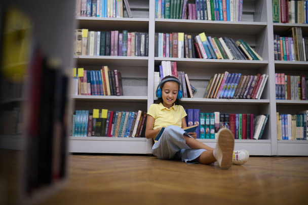 Modern schoolchildren. Girl in yellow tshirt sitting on floor in the library and listening to audio file - Фото, зображення