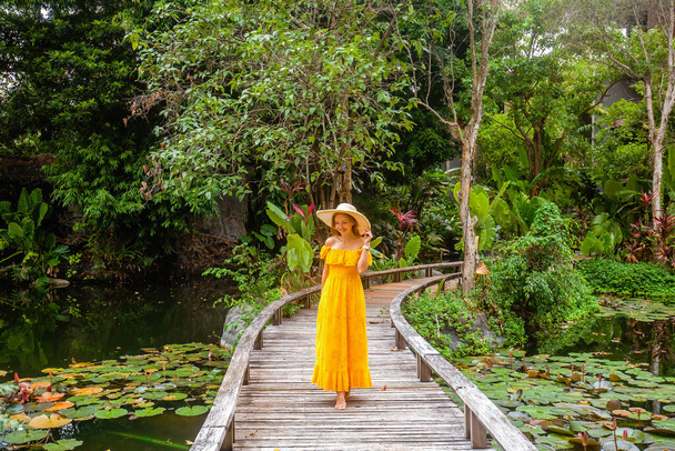 Happy woman in yellow dress enjoy her summer vacation in resort. Female traveller walking on wooden bridge among tropical greenery and green pond with water lilies in hot sunny day. Holiday and travel - Фото, зображення