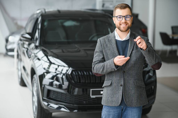 Portrait of a handsome salesman in the suit standing near the car in the showroom. - Zdjęcie, obraz