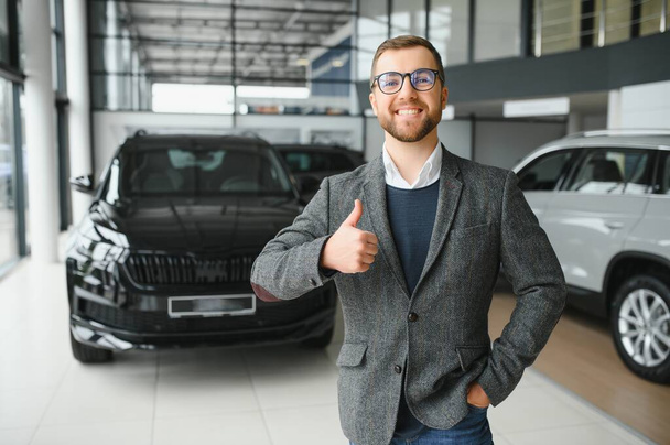 Portrait of a handsome salesman in the suit standing near the car in the showroom. - Foto, Imagem