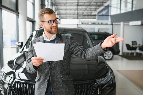 Portrait of a handsome salesman in the suit standing near the car in the showroom. - Foto, Imagen
