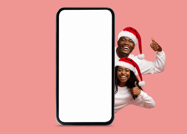 Щаслива чорна жінка і чоловік у капелюхах Санта-Клауса показують пальці на великих смартфонах з порожнім екраном, рекомендуйте додаток, ізольований на рожевому фоні. Пропозиція, реклама та додаток на Різдво, Новий рік - Фото, зображення