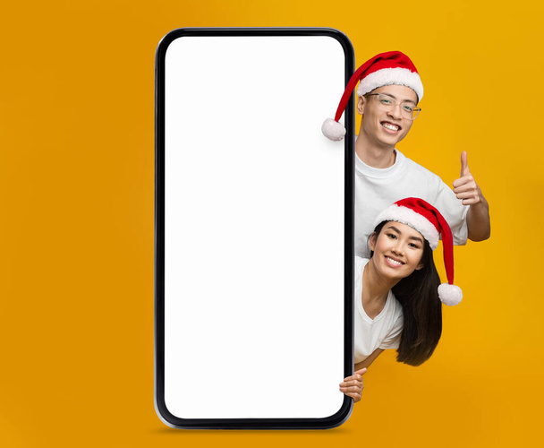 Щаслива молода жінка і хлопець з капелюхів Санта-Клауса показують великий палець з великим смартфоном з чистим екраном ізольованим на жовтому тлі. Пропонуй, рекламуй і додай поради щодо свята, Нового року і Різдва. - Фото, зображення