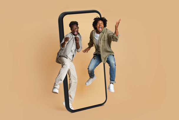 Szczęśliwi podekscytowani młodzi afrykańscy Amerykanie z otwartymi ustami radują się i skaczą po ogromnym ekranie smartfona, odizolowani na beżowym tle, studiu, pełnej długości. Aplikacja i gadżet dla zabawy z przyjacielem razem - Zdjęcie, obraz