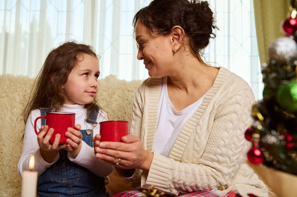 Beautiful multi-ethnic family of a loving mother and her lovely daughter, with mugs of hot chocolate drink, enjoying happy moments together at Christmas holidays in cozy home interior. December 25th - Photo, Image