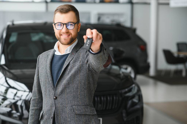 Portrait of a handsome salesman in the suit standing near the car in the showroom. - Foto, Imagem