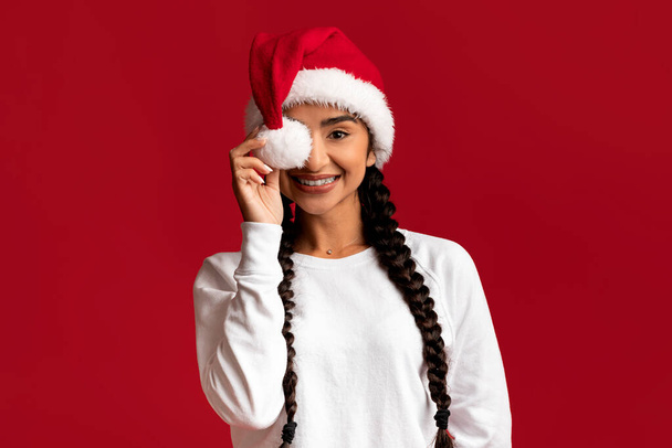 Playful Young Arab Female Wearing Santa Hat Covering Eye With Pompom And Looking At Camera, Happy Cheerful Middle Eastern Woman Enjoying Christmas Holidays, Posing Over Red Background, Copy Space - Photo, Image