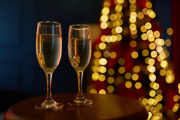 Close up of two glasses of champagne, sparkling wine standing on table. People celebrating new year in studio decorated with christmas tree. Concept of new year and holiday. - Foto, Imagem