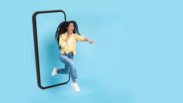 Yllättynyt innoissaan onnellinen arabi nainen hyppää ulos valtava älypuhelin näyttö, osoittaa sormella tyhjään tilaan, eristetty sinisellä taustalla, studio, panoraama. Suuri tarjous ja mainos, valtava myynti ja tunteet - Valokuva, kuva