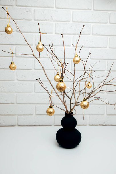 Рождественское украшение, сухие ветки деревьев в черной вазе украшены золотыми шариками против белой кирпичной стены. Вид спереди - Фото, изображение