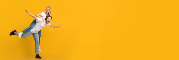 Gek aanbod. Vrolijk jong stel dat plezier heeft op gele achtergrond, brede horizontale banner met gelukkige Millennial echtgenoten die samen voor de gek houden, Man Piggybacking Woman, Extended Shot, Copy Space - Foto, afbeelding