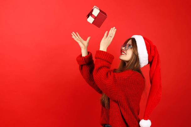 A young woman in a santa hat throws up a Christmas present on a red background, surprise. - Fotó, kép