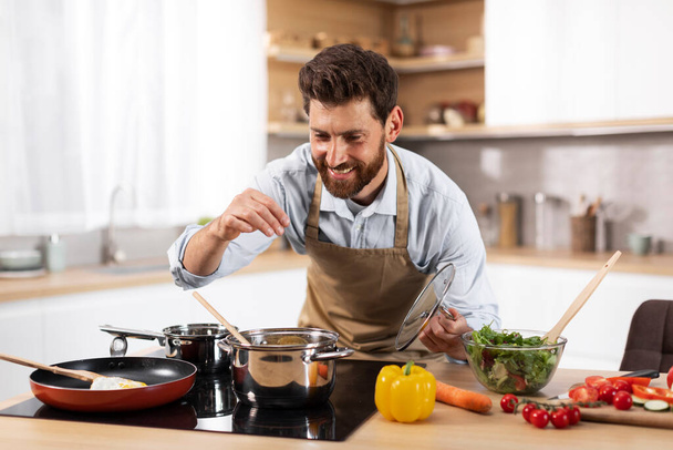 Cheerful adult european bearded man chef in apron prepares dinner for family, salts soup in modern kitchen interior at table with organic vegetables. Cook healthy meal at home, diet and food, covid-19 - Фото, изображение
