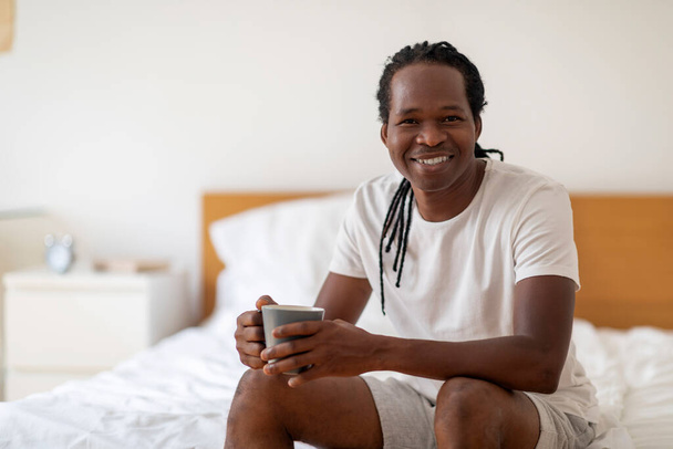 Knappe jonge zwarte man rustend op bed met kopje koffie in handen, Millennial Afro-Amerikaanse man genieten van warme drank en glimlachen op de camera terwijl ontspannen in gezellige slaapkamer thuis, kopiëren ruimte - Foto, afbeelding