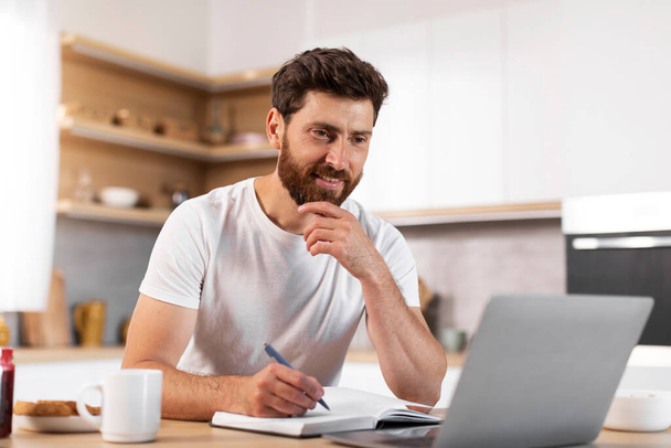 Happy handsome adult european bearded man making notes, watching video lesson on laptop in modern kitchen interior. Study, work remotely, video call and verbinar at home, new normal due covid-19 - Foto, Bild