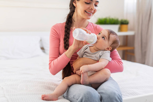 Baby care. Loving mom giving bottle with water or milk to her little daughter while relaxing together on bed in bedroom, copy space. Mother spending time with infant child at home - Photo, Image