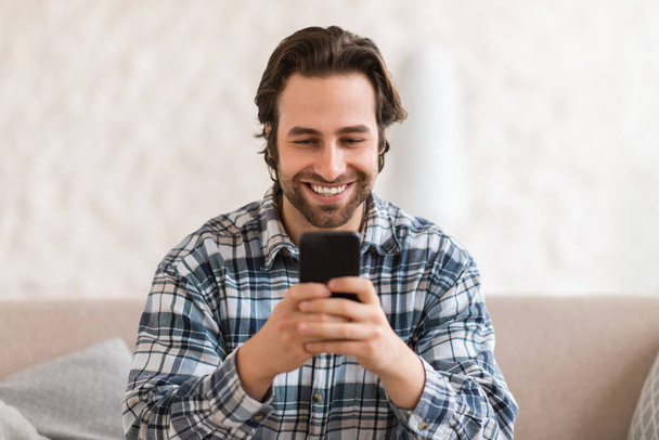 ソファの上で頑丈と国内服で幸せな千年の白人男性は、白い部屋のインテリアでスマートフォンでチャットを閉じます。新しいアプリ,デバイスでのリモート通信,ブログやソーシャルネットワーク - 写真・画像