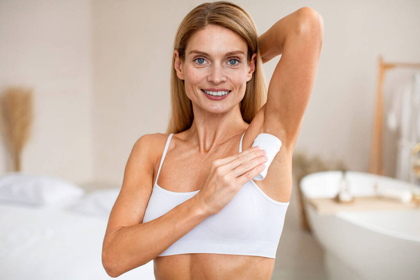 Portrait of positive middle aged woman using antiperspirant stick for underarms, applying deodorant to armpit for sweating prevention, standing in room interior - Photo, Image