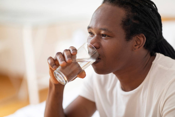 Hydratation corporelle. Jeune homme noir buvant de l'eau à partir de verre à la maison, gros plan portrait de beau Afro-Américain bénéficiant d'une boisson minérale saine tout en se relaxant dans la chambre, Copier l'espace - Photo, image