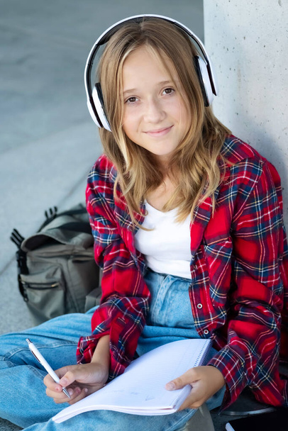 Молодий усміхнений красивий хіпстер щаслива дівчина-підліток на вулиці міста, прослуховування музики на навушниках, джинсах, рюкзаку, позитивний, весняний тренд, міський підліток стиль, ноти написання
. - Фото, зображення