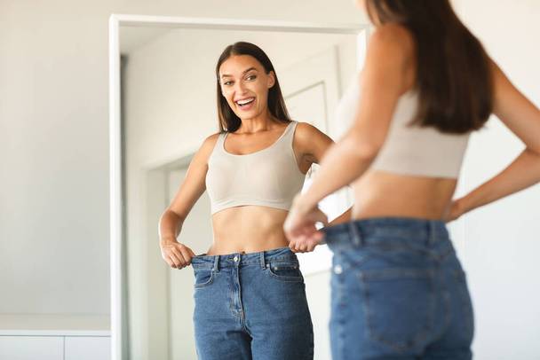 Успешная концепция похудения. Возбужденная кавказка в джинсах после потери веса, сравнивает размер, позирует возле зеркала дома, копирует пространство - Фото, изображение