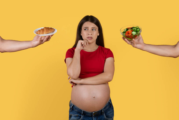 Важкий вибір. Сумнівна вагітна жінка вибирає між салатом і хорватом, дві руки дають їжу зі здоровою і нездоровою їжею завагітнілій леді, жовте тло, колаж - Фото, зображення
