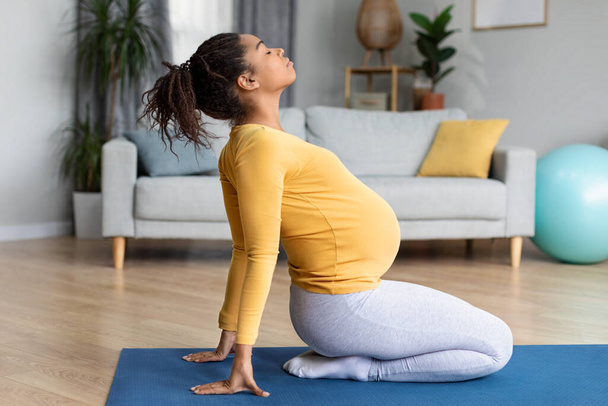 Happy pretty young black pregnant female with big belly doing exercises, practicing yoga in living room interior. Stretching training, body and health care, sports at home alone and expecting child - Foto, immagini