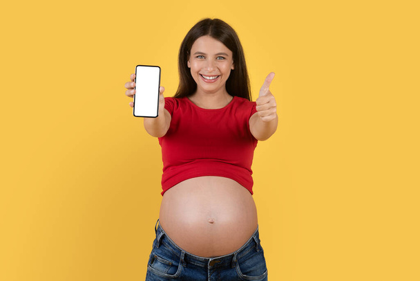いいアプリだ。空白のスマートフォンを表示し、親指をジェスチャー妊婦笑顔,幸せな期待の女性は、新しいモバイルアプリケーションをお勧めします,黄色の背景に孤立立ちます,モックアップ - 写真・画像