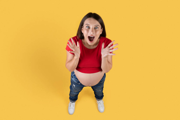 Above Shot Of Surprised Young Pregnant Woman Opening Mouth In Amazement, Joyful Expectant Female Looking At Camera, Emotionally Reacting To Nice Offer While Standing Isolated Over Yellow Background - Photo, Image