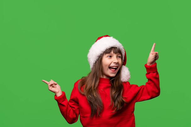 A cheerful smiling girl in a Santa hat shows with her index fingers discounts, promotions, gifts. Emotional portrait isolated on green background. New Year and Christmas concept. High quality photo - Фото, зображення