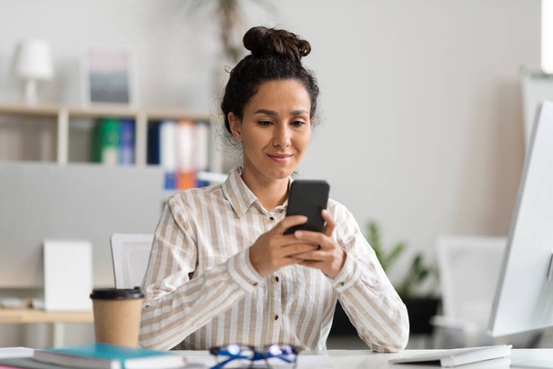 Усміхнена молода жінка офісний працівник використовує смартфон і читає повідомлення, сидячи за столом з ПК в офісному інтер'єрі. Робота професіонала, дизайнера з сучасними технологіями
 - Фото, зображення