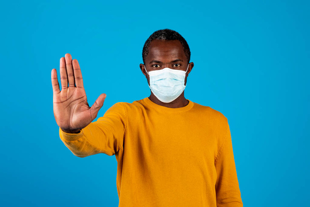 Tyytymätön afrikkalainen keski-ikäinen mies, jolla on sepelvaltimotaudin suojaava naamio, pitää kädestä stop-merkkiä, varoitus COVID-19: ltä, katsoen kameraa huolestuneella ilmeellä, sininen tausta - Valokuva, kuva