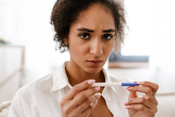 Genç siyahi kadın hamilelik testini evdeki kanepede oturarak yaparken üzülüyor. - Fotoğraf, Görsel