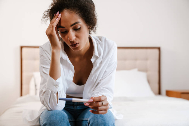 Young black woman being upset while holding pregnancy test sitting in bed at home - Foto, afbeelding
