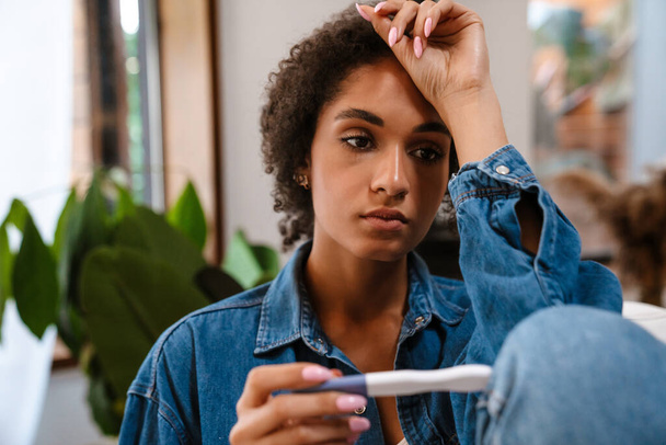 Young black woman being upset while holding pregnancy test sitting on sofa at home - Foto, imagen