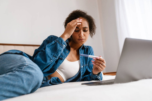 Young black woman holding pregnancy test and using laptop while lying in bed at home - Zdjęcie, obraz