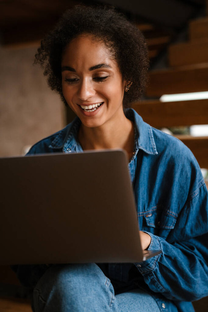 Young black woman wearing denim shirt smiling and using laptop at home - 写真・画像