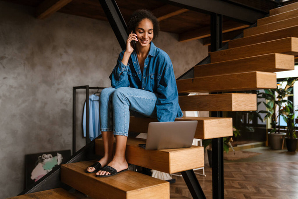 Young black woman wearing denim shirt smiling while using laptop and cellphone at home - Fotoğraf, Görsel