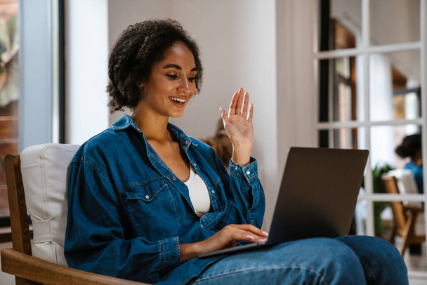 Young black woman wearing denim shirt smiling and using laptop at home - Zdjęcie, obraz