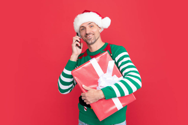 elfe joyeux parlant au téléphone. homme en santa claus chapeau salutation en ligne sur le téléphone avec boîte cadeau sur fond rouge. nouvel an. - Photo, image