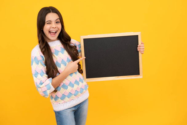 School sales board. Cheerful teenage girl kid hold blackboard chalkboard with copy space on yellow background. Excited face. Amazed expression, cheerful and glad - Photo, Image