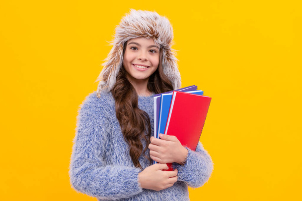 cheerful child in sweater and earflap hat with workbook on yellow background, education. - Fotó, kép