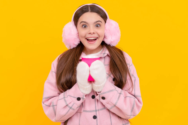 happy female fashion model on yellow background. teen girl in mittens with coffee cup. child wearing warm clothes. drinking tea. kid in fur earmuffs. - Photo, Image
