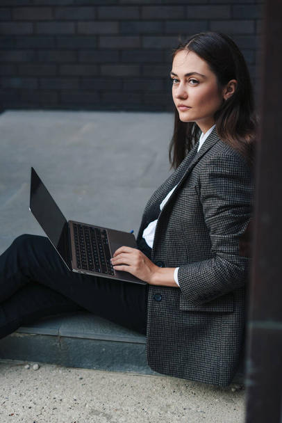Hermosa mujer de negocios con laptop y documentos, sentada en el piso apoyada en la pared del edificio de oficinas, mirando hacia otro lado. Gente corporativa. Hermosa. - Foto, imagen