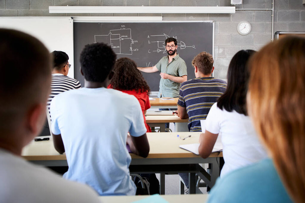 Male Tutor Teaching University Students In Classroom. Rear view from college attending at teacher in class. Professor explains the lesson on the blackboard. High quality photo - Zdjęcie, obraz