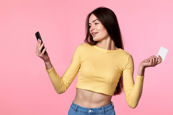 happy attractive woman in yellow clothes using mobile phone and plastic debit credit card isolated on pink background. Online payment - Fotó, kép